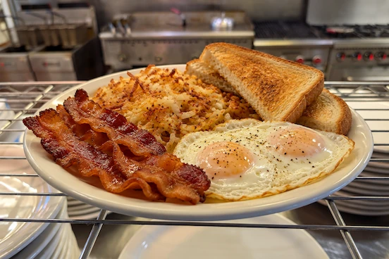 Two egg breakfast at Miller's Tavern with over-easy eggs, bacon, hash browns, and toast served on a white plate at the kitchen line.
